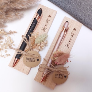 Könnte beinhalten: Zwei Stifte, einer schwarz und einer roségold, präsentiert auf Holzplatten. Jeder Stift ist mit einem kleinen Blumenstrauß und einem Anhänger mit der Aufschrift "DANKE" versehen. Die Holzplatten tragen den Text "Lehrerin mit".