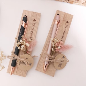 May include: Two pens, one black and one pink, with rose gold accents, presented on wooden cards. Each card has dried flowers and a tag that says "DANKE" and "Hebamme mit". The pens are tied with twine.