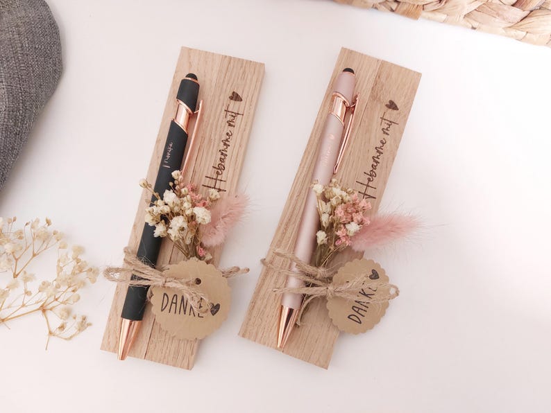 May include: Two pens, one black and one pink, with rose gold accents, presented on wooden cards. Each card has dried flowers and a tag that says "DANKE" and "Hebamme mit". The pens are tied with twine.