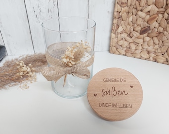 Könnte beinhalten: Ein rundes Holzschild mit dem Text "Genieße die süßen Dinge im Leben" und zwei kleinen Herzen. Das Schild liegt auf einer weißen Oberfläche neben einem Glasgefäß mit getrockneten Blumen und einem braunen Band.