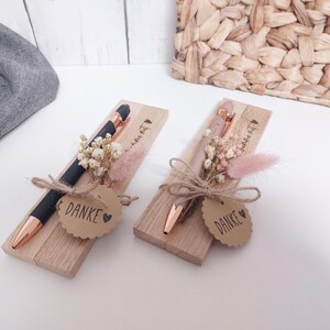 May include: Two pens in wooden boxes, decorated with dried flowers and a "Danke" tag. One pen is black with rose gold accents, the other is rose gold. The boxes are tied with twine.
