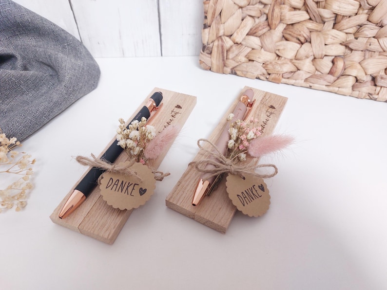 May include: Two pens in wooden boxes, decorated with dried flowers and a "Danke" tag. One pen is black with rose gold accents, the other is rose gold. The boxes are tied with twine.