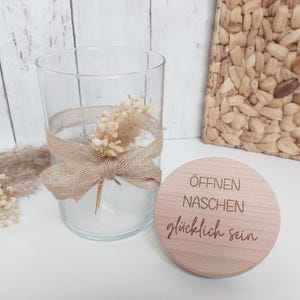 Könnte beinhalten: Ein Holzdeckel mit dem Text "Öffnen Naschen glücklich sein" in hellbrauner Schrift. Der Deckel liegt auf einer weißen Oberfläche neben einem durchsichtigen Glasgefäß mit einem braunen Band und getrockneten weißen Blumen.