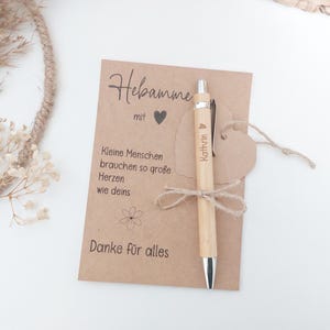 Könnte beinhalten: Ein hellfarbener Holzstift mit silbernen Akzenten ist mit einer Schnur an einer Kraftpapierkarte befestigt. Die Karte hat deutschen Text und ein Herzsymbol. Der Stift ist mit einem Namen graviert. Die Karte ist mit einer kleinen Blume verziert.