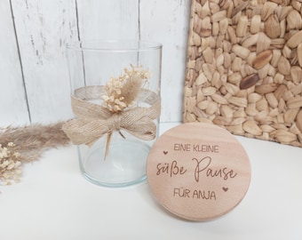 Könnte beinhalten: Ein Holzdeckel mit dem Text "Eine kleine süße Pause für Anja" eingraviert. Der Deckel liegt auf einer weißen Oberfläche neben einem Glasgefäß mit getrockneten Blumen und einem braunen Band.