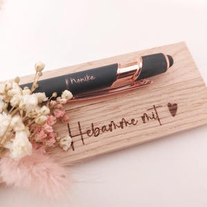 May include: A black pen with rose gold accents and the name "Monika" engraved on it, resting on a wooden plaque. The plaque has the words "Hebamme mit" and a heart symbol engraved. A small bouquet of dried flowers is placed near the pen.