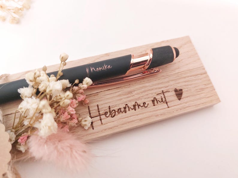 May include: A black pen with rose gold accents and the name "Monika" engraved on it, resting on a wooden plaque. The plaque has the words "Hebamme mit" and a heart symbol engraved. A small bouquet of dried flowers is placed near the pen.