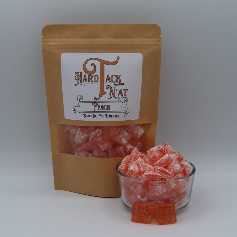 May include: A brown paper bag with a label that says "Hardtack N'at" and "Peach" with the tagline "Tastes Like You Remember". The bag is filled with pink hard candies. A small glass bowl filled with the same pink hard candies is in front of the bag.