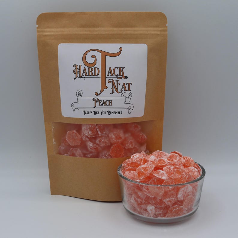 May include: A brown paper bag with a label that reads "Hard Tack N'at Peach" and a clear window showing peach flavored hard candies. The candies are in a glass bowl and are a light orange color.