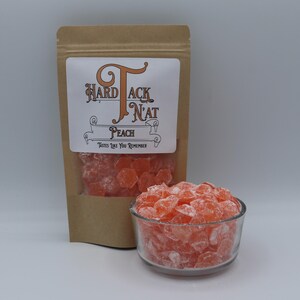 May include: A brown paper bag with a white label that says "Hard Tack N'at" and "Peach" with the tagline "Tastes Like You Remember". The bag contains peach flavored hard candies. A small glass bowl filled with the candies is in the foreground.