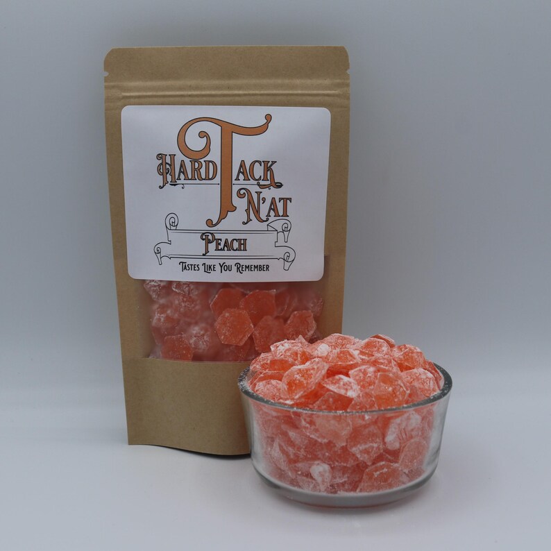 May include: A brown paper bag with a white label that says "Hard Tack N'at" and "Peach" with the tagline "Tastes Like You Remember". The bag contains peach flavored hard candies. A small glass bowl filled with the candies is in the foreground.