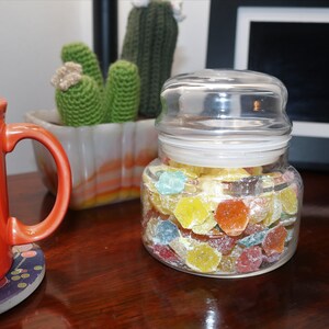 May include: A glass jar filled with colorful, sugar-coated candies. The candies are in various shapes and sizes, including stars, hearts, and circles. The jar has a clear lid and is sitting on a wooden surface.