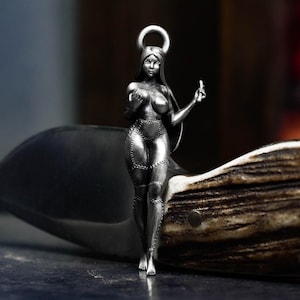 May include: A silver-colored pendant depicting a stylized female figure with a halo. The figure is wearing a chainmail-like outfit and is holding a heart in one hand and making a gesture with the other. The pendant is resting on a brown and white surface.