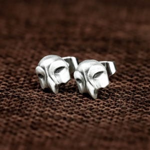 May include: A pair of silver stud earrings, each shaped like a mask with a pointed beak and eye slits. The earrings are presented on a textured brown surface.