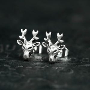 May include: A pair of silver stud earrings, each shaped like a deer head with detailed antlers. The earrings are presented on a dark, textured surface, accentuating their metallic shine. The earrings are made of silver.