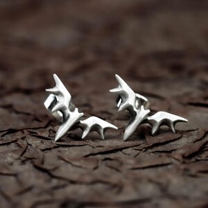 May include: A pair of silver-colored bat-shaped stud earrings. Each earring features a detailed design with pointed ears and wing details. The earrings are displayed on a textured, brown surface.