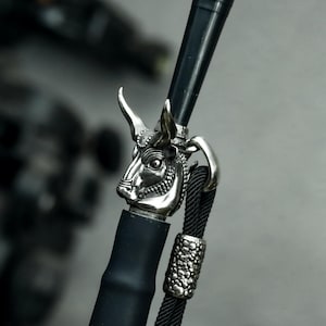May include: A black walking stick with a silver bull head design. The bull head is detailed with horns and facial features. A black rope and decorative silver band are attached to the stick.