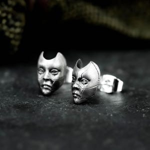 May include: A pair of silver-toned stud earrings. Each earring is sculpted in the shape of a stylized human face, with detailed features and a mechanical or futuristic aesthetic. The earrings are set against a dark background.