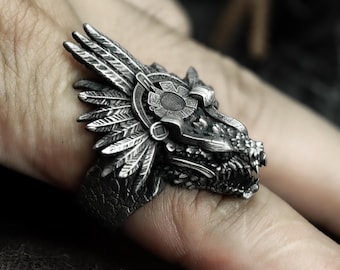May include: A detailed silver ring featuring a dragon head design. The ring showcases intricate details, including feathers, a circular emblem, and a textured surface. The dragon's mouth is open, revealing sharp teeth. The ring is worn on a finger.