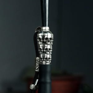 May include: A black and silver hookah stem with a stacked skull design. The stem has a black handle and a silver metal skull design. A black rope is attached to the stem.