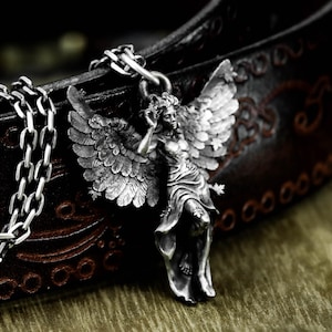 May include: A silver-colored pendant necklace featuring a detailed angel figure with outstretched wings. The angel is depicted in a flowing gown, with intricate feather detailing on the wings. The necklace chain is a chunky, silver-toned link. The background is a dark brown leather.