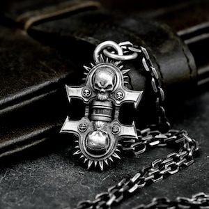 May include: Silver-toned pendant necklace featuring a detailed skull design. The pendant has a cross-like shape with multiple skulls and a chain link necklace. The necklace is displayed on a dark surface.