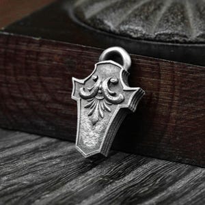 May include: A silver-colored pendant with an ornate, shield-like design. The pendant features a detailed floral and scrollwork pattern, with a loop at the top for a chain. The background is a dark wooden surface.
