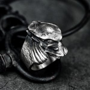 May include: A detailed, silver-coloured ring featuring a sculpted alien face with intricate details. The ring's design includes a helmet-like structure and a textured surface, set against a dark, blurred background. The ring is a statement piece.