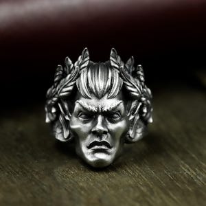 May include: A silver-toned ring featuring a detailed face with a stern expression, framed by a laurel wreath. The ring has a vintage, antique appearance, with intricate detailing on the face and the surrounding leaves.