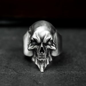 May include: A silver skull ring with detailed facial features, including a prominent brow, eye sockets, and teeth. The ring is set against a dark background, highlighting the metallic sheen and intricate design of the skull.