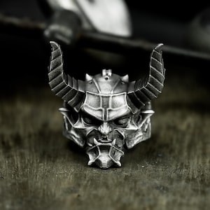 Op de afbeelding: Een zilverkleurige ring met een gedetailleerd demonisch gezicht en grote, gebogen hoorns. De ring heeft ingewikkelde texturen en een donkere, verouderde uitstraling, wat wijst op een gotisch of fantasiethema. De ring is ontworpen om aan een vinger te worden gedragen.