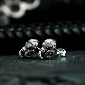 May include: A pair of silver-toned stud earrings, each shaped like a miniature gas mask. The earrings have detailed features, including circular filters and a textured surface. The design is industrial and edgy.