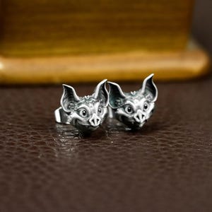 May include: A pair of silver-colored bat-shaped rings. Each ring features a detailed bat head with large ears, a small nose, and expressive eyes. The rings are set against a brown background, highlighting the intricate design and metallic sheen.