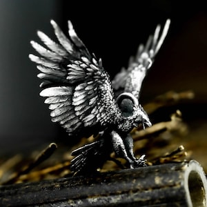 May include: A detailed silver-colored sculpture of a bird with outstretched wings. The bird is perched on a dark, textured surface, with its wings and feathers intricately carved. The sculpture has a dark background.