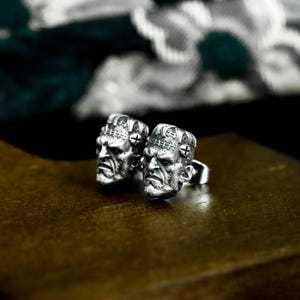 May include: A pair of silver-colored Frankenstein monster-themed rings. Each ring features a detailed sculpted face with stitched scars and bolts. The rings are displayed on a dark brown surface, with a blurred background.