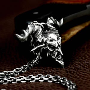 May include: A silver-colored pendant necklace featuring a detailed, demonic face with horns and sharp teeth. The pendant is attached to a chain with a series of interlocking links. The background is dark, highlighting the intricate details of the pendant.
