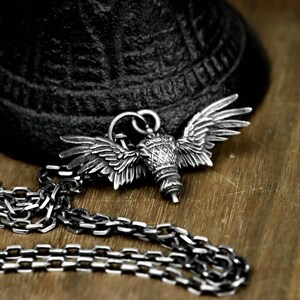Könnte beinhalten: Eine silberfarbene Halskette mit einem Anhänger in Form von Flügeln. Der Anhänger hat detaillierte Flügel und ein zylindrisches Zentralelement. Die Halskette hat ebenfalls eine silberne Kette mit einem Kettendesign. Der Hintergrund umfasst ein strukturiertes schwarzes Objekt und eine Holzoberfläche.