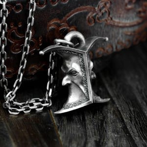 Puede incluir: Collar con colgante plateado que presenta un rostro estilizado con nariz puntiaguda y tocado curvo. El colgante está unido a una cadena plateada. El fondo es una superficie oscura y texturizada.