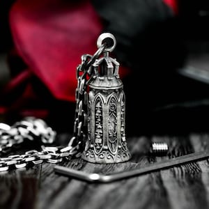 May include: A silver-colored pendant necklace featuring a bell-shaped charm with gothic architectural details and skull motifs. The pendant hangs from a chain, with a small screw and a metal tool visible nearby. The background is dark with a red accent.