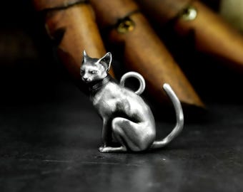 Puede incluir: Colgante de gato plateado en posición sentada, con un lazo para collar. El gato tiene collar y cola curvada. El fondo es oscuro, con elementos de madera borrosos.