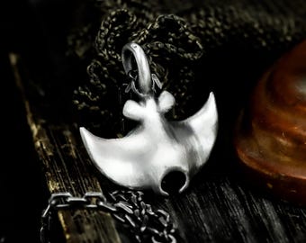 May include: A silver-colored pendant in the shape of a manta ray, suspended from a dark chain. The pendant has a brushed metal finish and is set against a dark, textured background. The design features curved wings and a detailed head.