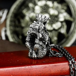May include: A detailed silver pendant necklace featuring an Aztec warrior design. The pendant showcases intricate carvings of a warrior figure with a feathered headdress and detailed armor. The necklace has a dark chain.