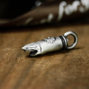 May include: A silver-colored pendant shaped like a bullet with a shark head design. The shark's mouth is open, revealing sharp teeth. The pendant has a loop at the end for a chain. The background is a blurred wooden surface.