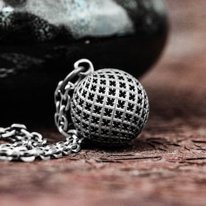 May include: A silver pendant necklace with a sphere-shaped charm. The sphere has a lattice pattern, and hangs from a chain. The background is a blurred dark shape on a textured, brown surface.