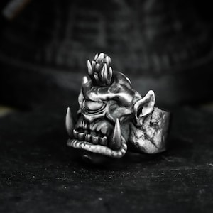 May include: A silver-colored ring featuring a detailed orc face design. The ring has a textured surface and prominent tusks, with a stylized crown on top of the head. The design is highly detailed, with visible wrinkles and facial features.