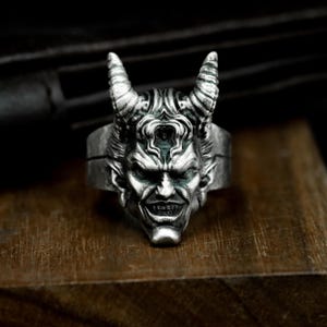 May include: A silver-colored ring featuring a detailed devil face with horns, a sinister grin, and intricate facial details. The ring is set against a dark background, highlighting the gothic and edgy design. The ring is a statement piece.