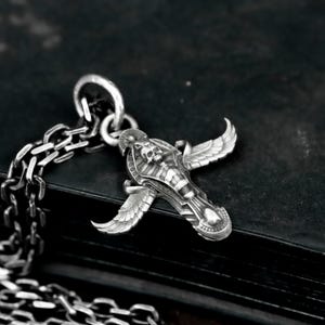 Może przedstawiać: Srebrny naszyjnik z wisiorkiem w kształcie stylizowanego sarkofagu ze skrzydłami. Wisiorek z czaszką zawieszony jest na grubym łańcuchu. Ciemne tło podkreśla misterne detale biżuterii.