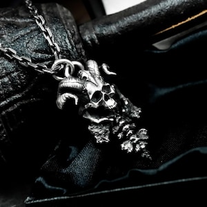 May include: A silver-toned pendant necklace featuring a skull with curved horns and a textured, detailed design. The skull pendant is attached to a chain necklace. The background is dark, emphasizing the gothic style of the jewelry.