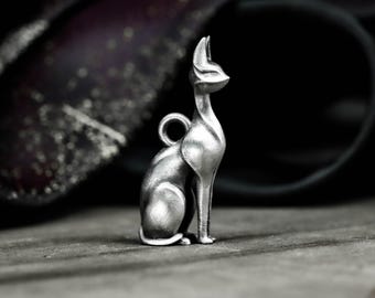 Puede incluir: Un colgante de plata con forma de gato sentado, con un lazo para colgar. El gato tiene un cuerpo esbelto, orejas puntiagudas y mira hacia un lado. El fondo es oscuro y borroso, con un indicio de superficie texturizada.