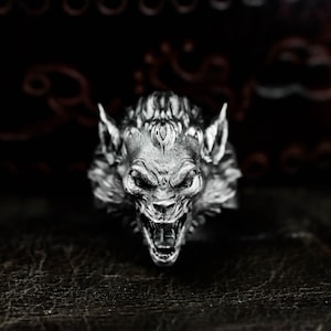 Puede incluir: Anillo plateado que representa una cabeza de hombre lobo con la boca abierta, dientes afilados y orejas puntiagudas. El anillo tiene una superficie texturizada y está sobre un fondo oscuro, resaltando el diseño detallado.